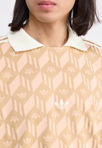 Peach-toned polo shirt with a diamond pattern and white Adidas logo. Features a contrasting white collar and fabric texture.