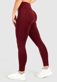 Bordeaux leggings met een hoge tailleband, naadloos ontwerp, textuuraccenten aan de achterkant en een aansluitende pasvorm, gedragen met witte sneakers.