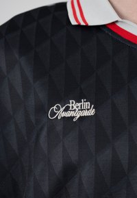 Camisa negra con un sutil patrón de diamantes, cuello a rayas blancas y rojas, y texto bordado en blanco "Berlin Avantgarde" en el pecho.