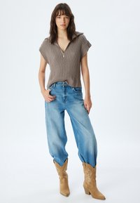 Brauner gerippter Pullover mit kurzem Ärmel und Reißverschlusskragen, kombiniert mit hellblauen übergroßen Jeans und hellbraunen Cowboy-Stiefeln mit Stickdetails.
