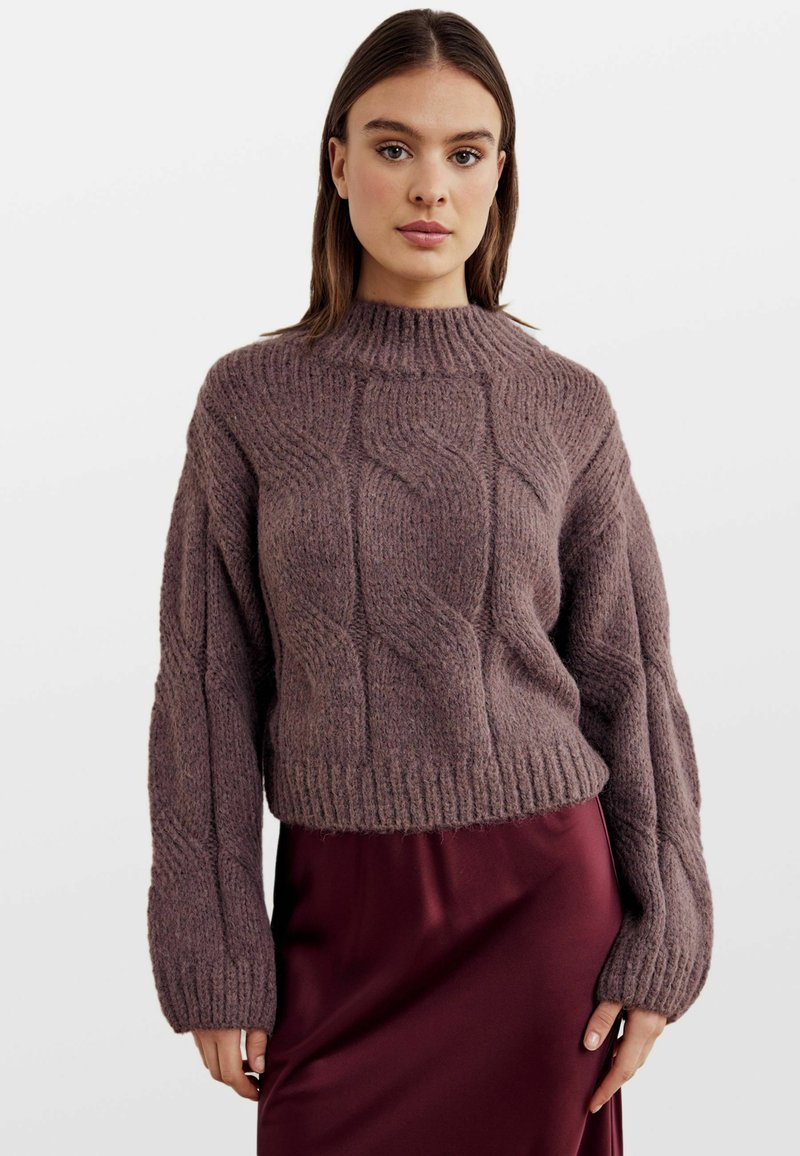 Maglione chunky a trecce in morbido mauve tenue. Presenta un collo alto e maniche larghe e oversize. Abbinato a una gonna in satin bordeaux.