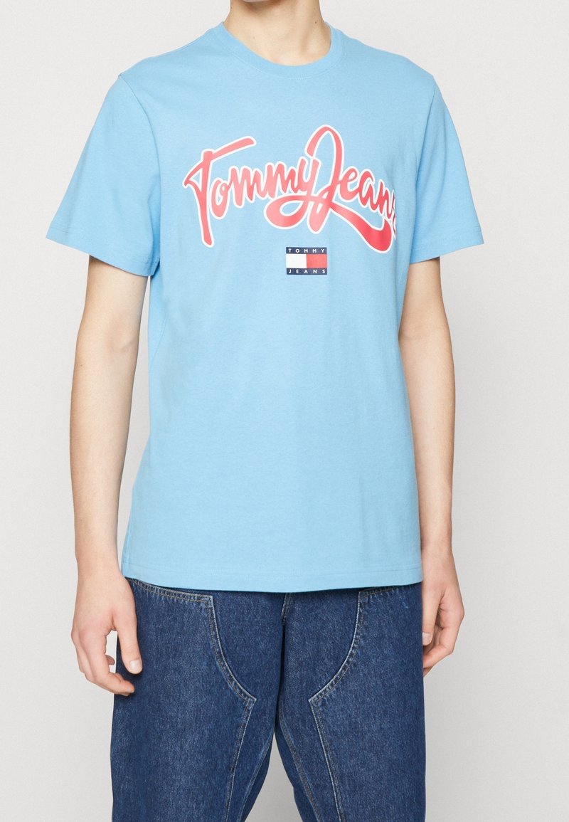 Light blue short-sleeve T-shirt with "Tommy Jeans" in large red script and logo, worn with loose dark blue jeans.