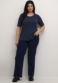 Camiseta de color azul marino, de manga corta y cuello redondo, combinada con jeans acampanados de color azul oscuro. La modelo se encuentra de pie con una mano en el bolsillo.