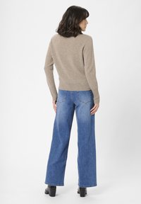 Kvinde med skulderlangt mørkt hår iført en beige sweater, blå vidde jeans og sorte hæle boots, vender væk fra kameraet mod en enkelt baggrund.