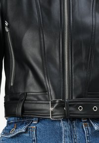 Svart skinnbikerjakke med sølvfargede glidelåser og belte i midjen. Har horisontale sømmer og metall hardwaredetaljer. Denim jeans er synlige nedenfor.