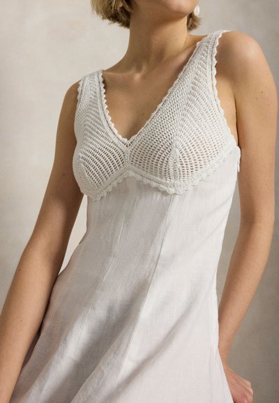 Sieviete valkā baltu bezroku kleitu ar tamborētu augšdaļu un V veida izgriezumu, uz neitrāla fona.