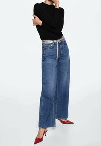 Jean en denim bleu à jambe large et taille haute, avec des coutures détaillées et une ceinture en chaîne argentée. Assorti avec un top noir à manches longues et des talons rouges.