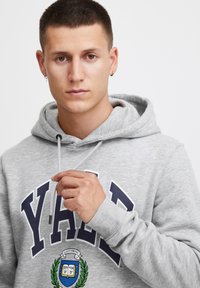 Sivá mikina s kapucňou s veľkým potlačou "YALE" v námořníckej farbe a logom štítu s vavrínovými akcentmi. Nastaviteľná šnúrka na kapucni.
