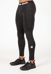 Zwarte sportleggings met een strakke pasvorm, een brede tailleband met witte tekst en een klein logo op de onderkant van het been. Matte textuur.