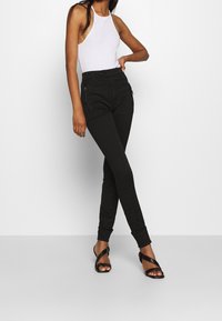 Pantalones negros de talle alto con un diseño ajustado, bolsillos con cremallera y puños enrollados, combinados con una camiseta sin mangas blanca y tacones negros con tiras.