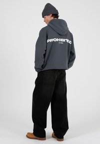 Felpa grigia con la scritta "PROHIBITED" in bianco sul retro, abbinata a jeans neri larghi e scarpe slip-on color cuoio. Berretto lavorato a maglia in testa.