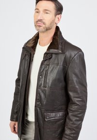 Man in een donkerbruine leren jas met een bontkraag boven een witte trui, staand tegen een effen lichte achtergrond.