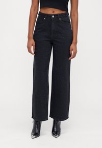 Calças de ganga largas pretas com embellishments de tachas, cintura alta e um design clássico de cinco bolsos, combinadas com botas pretas de bico fino.