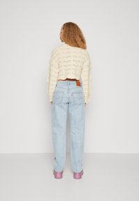 Cremfarbener gestrickter, kürzerer Pullover mit offener Maschenweise, kombiniert mit hellblauen geraden Jeans und pinken Schuhen. Rückansicht.