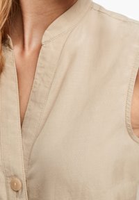 Beige, ärmlös topp med v-ringning och en enda knäppning. Tillverkad av ett mjukt, lätt tyg med en subtil textur.