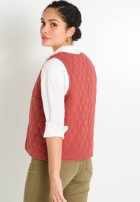 Gilet rouge matelassé avec un motif en losange, design sans manches, encolure ronde et coupe décontractée, porté sur une chemise à manches longues blanche.