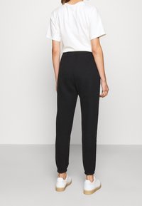 Kvinna klädd i svarta joggarbyxor, vit t-shirt och vita sneakers som står vänd bortåt mot en enkel bakgrund.