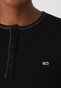 Sort ribbet henley-top med rund halsudskæring, gyldne sting, og fire sorte knapper, samt et lille broderet logo på brystet.