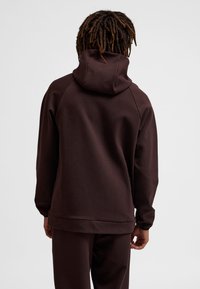 Mężczyzna ubrany w ciemnobrązową bluzę z kapturem i dopasowane spodnie, widziany od tyłu na jednolitym jasnym tle.