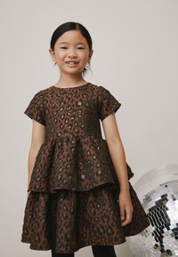 Barna leopárdmintás ruha rövid ujjakkal, réteges fodros kialakítással, texturált anyagból, arany díszítéssel. Alatta fekete leggings.