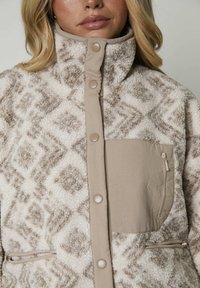 Giacca in fleece beige e crema con motivo a rombi, colletto alto, bottoni a scatto e tasche con cerniera. Tessuto testurizzato con accenti morbidi.