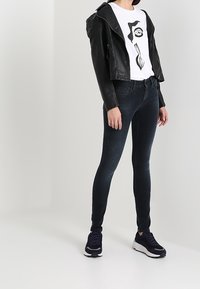 Musta lyhyt nahkatakki valkoisen paidan päällä, jossa on abstrakti silmäkuvio, yhdistetty tummiin kapeisiin farkkuihin ja tummansinisiin urheilukenkiin.