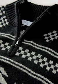 Gestrickter schwarz-weißer Zip-Pullover mit hohem Kragen, der ein geometrisches Muster aus Karos und Streifen aufweist, sowie einen metallischen Reißverschluss.
