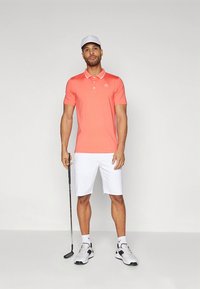 Korálové polo tričko s bielym golierom, v kombinácii s bielymi šortkami. Model drží golfovú palicu, má na hlave sivú čiapku a biele tenisky s akcentmi.