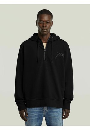 Jeune homme portant un sweat à capuche noir zippé avec une capuche et un jean bleu clair, debout devant un fond clair uni.