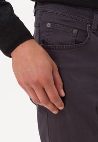 Mano maschile appoggiata all'anca vicino alla tasca di pantaloni grigio scuro e manica di maglione nero, che mostra le cuciture e i rivetti metallici sulla tasca.