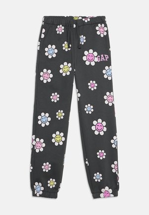 Pantalon de survêtement gris foncé avec ceinture et poignets élastiques, couvert de fleurs blanches souriantes aux visages pastel rose, bleu et jaune, avec le logo "GAP".