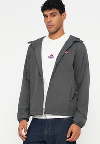 Szara lekka kurtka z kapturem, elastycznymi mankietami, zapięciem na zamek i małym logo na piersi, noszona na białej koszulce i niebieskich dżinsach.