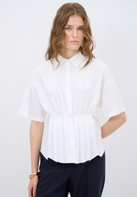 Camisa blanca de cuello con mangas cortas, con un diseño plisado en la cintura y una textura suave. Combinada con pantalones oscuros.