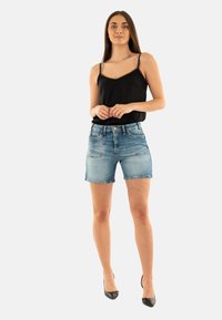 Top senza maniche nero con spalline di perline, abbinato a pantaloni corti di jeans blu chiaro. La modella indossa tacchi alti neri, in piedi contro uno sfondo bianco.