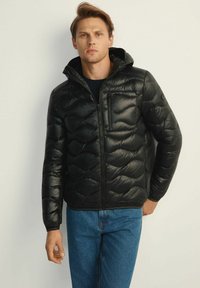 Czarna kurtka puchowa o błyszczącym wykończeniu, z zapięciem na zamek błyskawiczny z przodu, kapturem i ukośnym wzorem pikowania. Noszona z niebieskimi dżinsami.