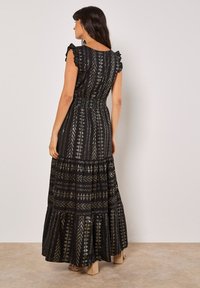 Robe maxi noire avec des motifs géométriques dorés métalliques, taille smockée, manches volantées et ourlet à niveaux. Texture de tissu lisse.