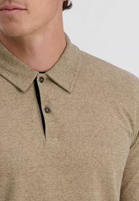 Mann trägt ein beiges Poloshirt aus strukturierter Stoffqualität mit zwei am Kragen befestigten Knöpfen vor einem einfarbigen Hintergrund.