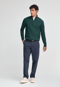 Mörkgrön zip-up tröja, vit innerlager, marinblå byxor, beige sneakers; slät textur, åtsittande design, minimal varumärkesdetalj.