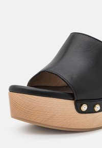 Sandale noire en cuir à bout ouvert avec semelle plateforme en bois et clous dorés ornés de petites perles blanches sur le côté.