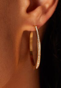 Gouden oorring met een gladde oppervlakte en een rij heldere kristallen aan één rand, tegen een getinte oor.
