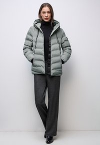 Ljusgrön vadderad jacka med huva, försedd med dragkedja framtill, sidofickor och ett quiltat mönster. Bärs över en svart polotröja.