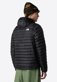 Chaquetón negro acolchado con capucha, que presenta una textura de acolchado y un pequeño logo en la parte superior izquierda. Combinado con pantalones de color verde claro.