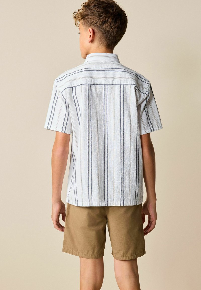 Dreng stående med ryggen mod, iført en kortærmet hvid skjorte med lodrette gråstriber og lysebrune shorts mod en ensfarvet baggrund.
