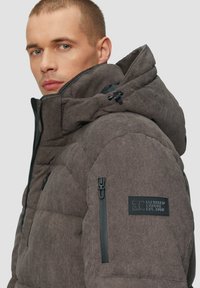 Veste matelassée grise avec un col montant, fermeture éclair noire et patch de marque. Elle présente un tissu texturé et des cordons de réglage pour la capuche.