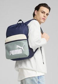 Zaino blu navy con parte superiore beige chiaro e frontale verde menta, con un grande logo bianco Puma. Realizzato in tessuto resistente.
