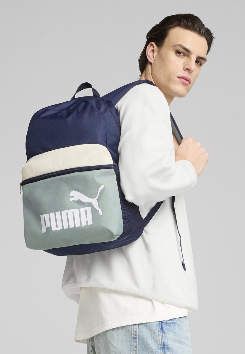 Zaino blu navy con parte superiore beige chiaro e frontale verde menta, con un grande logo bianco Puma. Realizzato in tessuto resistente.