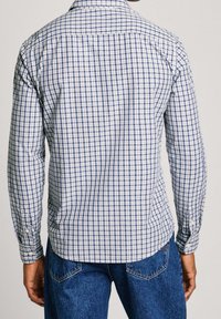 Uomo che indossa una camicia a maniche lunghe a scacchi blu e bianchi e jeans blu, visto di spalle su uno sfondo semplice.