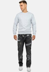 Felpa grigia con maniche lunghe, abbinata a jeans in denim neri caratterizzati da toppe ricamate e numerose tasche con zip. Sneakers bianche.