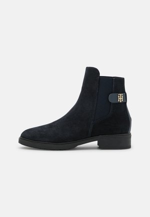 Bottine Chelsea en daim noir avec talon bas, panneau latéral élastique et détail logo doré sur le côté extérieur.