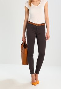 Tmavě hnědé přiléhavé kalhoty doplněné tanovým opaskem, bílým krátkým rukávem a žlutými špičatými podpatky. Modelka drží hnědou tašku.